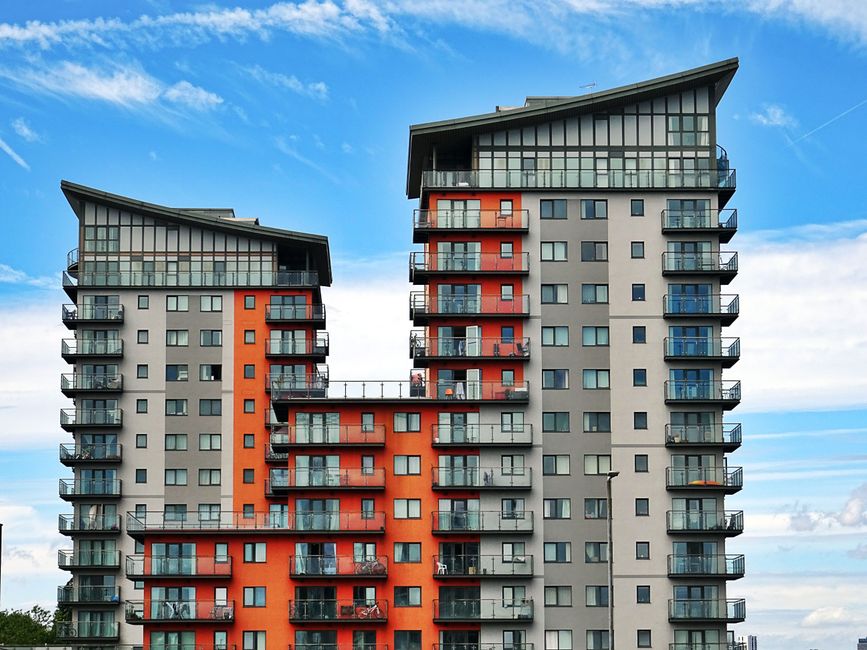 A high-rise residential building with a concrete façade featuring numerous rectangular windows arranged in vertical columns. Some windows are open, revealing interior curtains and furniture, while ot