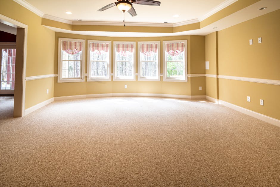 An empty, spacious living room with beige carpet flooring that appears freshly cleaned and free of dust or stains. The room features a large bay window with five sections, each fitted with white frame