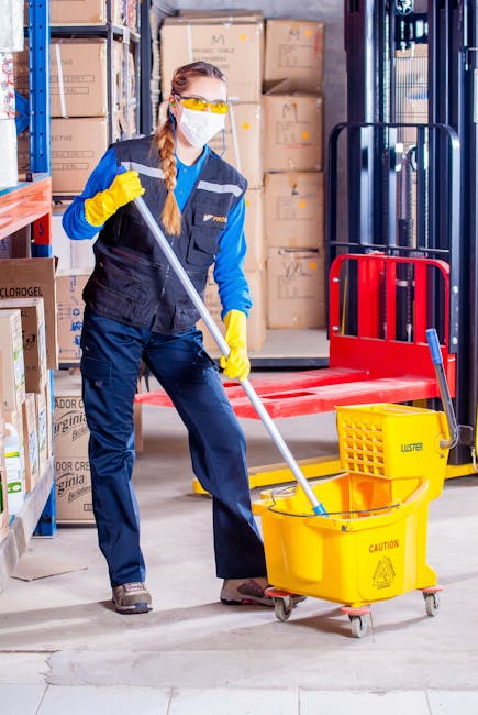 A commercial cleaning trolley positioned against a tiled wall in an office corridor, containing various cleaning tools and supplies. The trolley holds a vacuum cleaner, mop, bucket, spray bottles, clo
