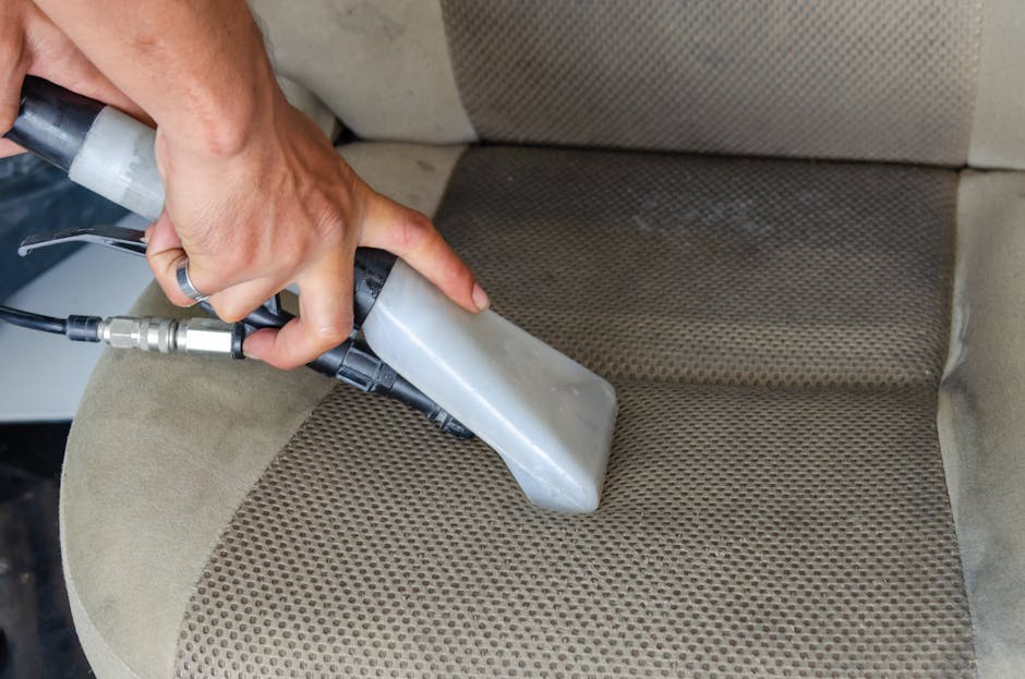 A person wearing a dark leather jacket is vacuuming a decorative yellow and white patterned cushion placed on a dark fabric sofa in a residential room, with the vacuum hose held in one hand and the so